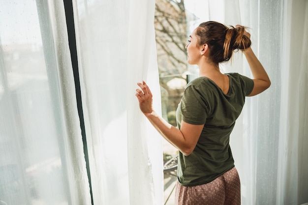 Jonge vrouw genieten van uitzicht door het raam in de ochtendzon thuis of hotelkamer.