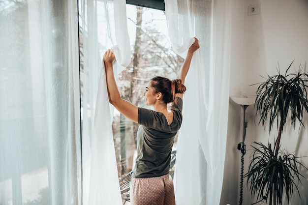 Jonge vrouw genieten van uitzicht door het raam in de ochtendzon thuis of hotelkamer.