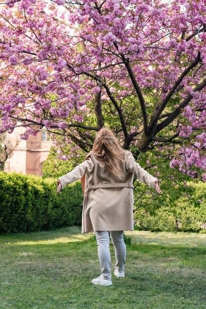 Jonge vrouw genieten van warme lentedag in park in de buurt van kersenbloesem Achteraanzicht portret van meisje in de buurt van bloeiende boom Verticaal frame