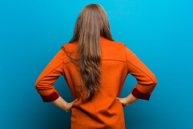 Jonge vrouw gevoel verward of vol of twijfels en vragen, benieuwd, met de handen op de heupen, achteraanzicht tegen blauwe muur