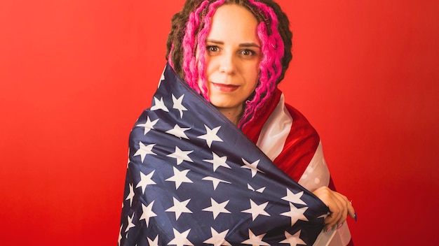 Jonge vrouw gewikkeld in Amerikaanse vlag op rode achtergrond Gelukkige vrouw glimlachend en kijkend naar camera poseren in studio Concept van Flag Day nationale feestdag