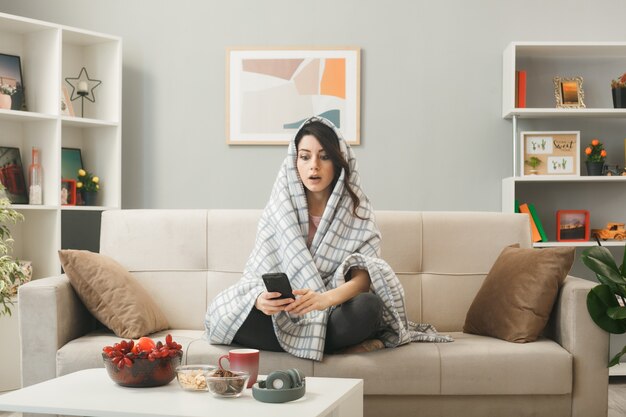 Jonge vrouw gewikkeld in een plaid die de telefoon vasthoudt en kijkt op de bank achter de salontafel in de woonkamer