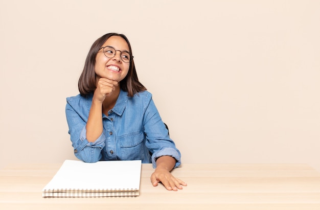 Jonge vrouw glimlachend met een gelukkige, zelfverzekerde uitdrukking met de hand op de kin, zich afvragend en opzij kijkend