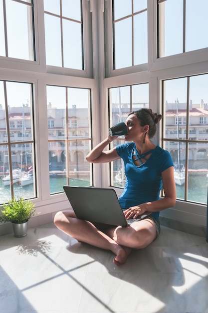 Jonge vrouw het drinken koffie, die met laptop bij het venster thuis over de jachthaven werken. F