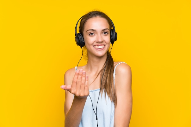 Jonge vrouw het luisteren muziek over geïsoleerde gele muur die met hand uitnodigt te komen. Blij dat je bent gekomen