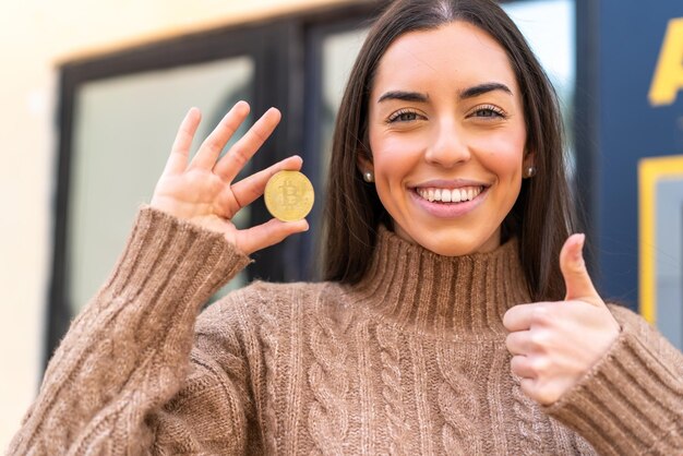 Foto jonge vrouw houdt een bitcoin buitenshuis vast met duimen omhoog omdat er iets goeds is gebeurd