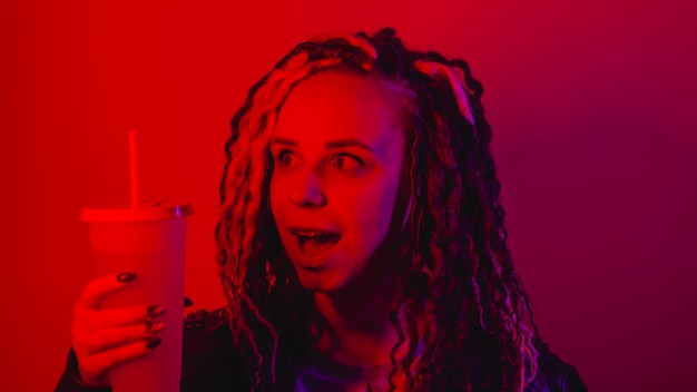 Jonge vrouw houdt grote kop frisdrank met stro in donkere kamer Portret van vrouw poseren met niet-alcoholische drank in duisternis verlichting van rood licht