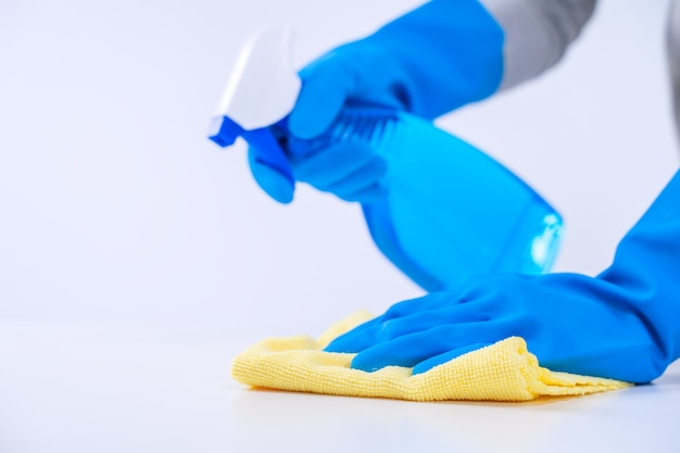 Jonge vrouw huishoudster doet het schoonmaken van witte tafel in schort met blauwe handschoenen, spray cleaner, natte gele lap, close-up, kopieer ruimte, blanco ontwerpconcept.