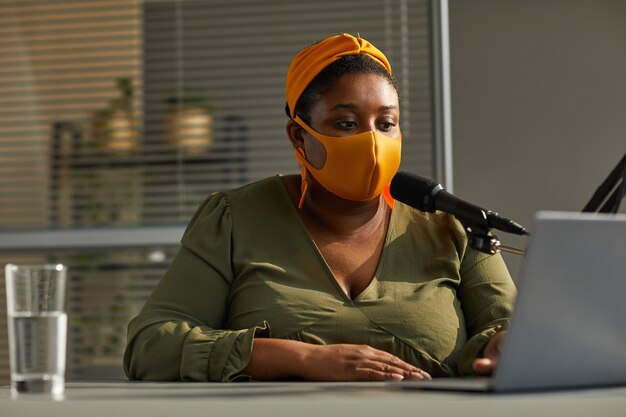 Jonge vrouw in beschermend masker spreken op microfoon en kijken op de monitor van laptop heeft ze online interview