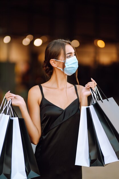 Jonge vrouw in beschermend steriel medisch masker op haar gezicht met boodschappentassen die tijdens het winkelen genieten.