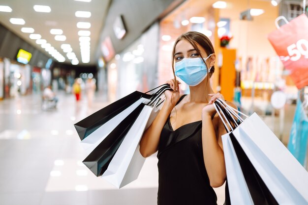 Jonge vrouw in beschermend steriel medisch masker op haar gezicht met boodschappentassen in het winkelcentrum.