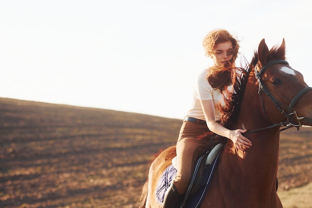 Jonge vrouw in beschermende hoed met haar paard in landbouwveld op zonnige dag