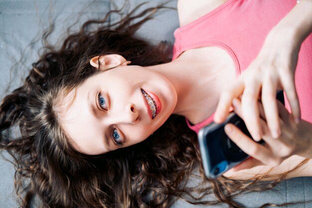 Foto jonge vrouw in beugels met lang krullend haar ligt op de bank en scrolt sociale netwerken op haar telefoon