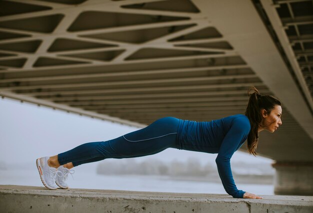 Jonge vrouw in blauw trainingspak die pushups doet onder de brug in de stedelijke omgeving