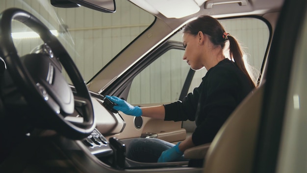 Jonge vrouw in blauwe handschoenen maakt het interieur van de auto schoon voor luxe SUV, close-up