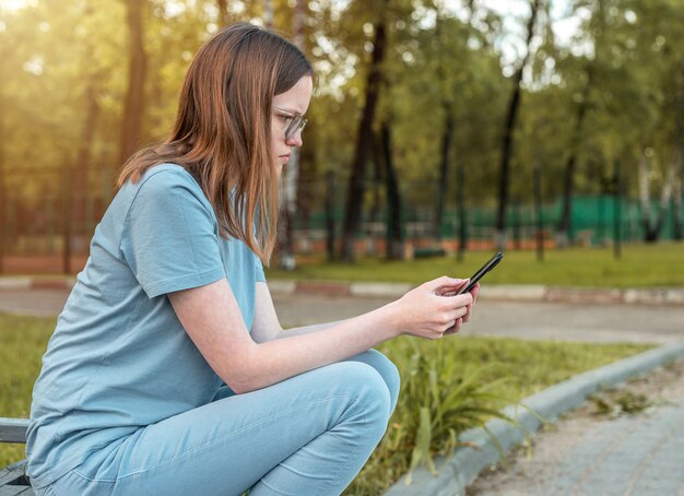 Jonge vrouw in brillen zittend op een bankje alleen en met behulp van mobiele telefoon persoon op zoek op smartphone in...