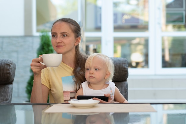 Jonge vrouw in café met kind in haar armen drinkt koffie en kijkt naar de telefoon. Moderne zakelijke moeder.