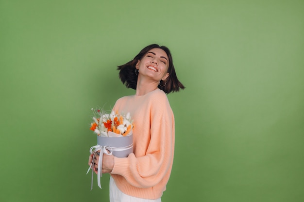 Jonge vrouw in casual perzik trui geïsoleerd op groene olijf muur houden oranje witte bloembak samenstelling van katoen bloemen gypsophila tarwe en lagurus voor een geschenk blij verbaasd verrast