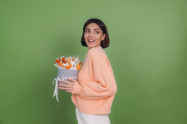 Jonge vrouw in casual perzik trui geïsoleerd op groene olijf muur houden oranje witte bloembak samenstelling van katoen bloemen gypsophila tarwe en lagurus voor een geschenk blij verbaasd verrast