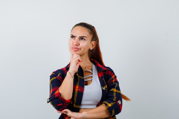 Jonge vrouw in crop top, geruit hemd staat in denkende pose en kijkt peinzend, vooraanzicht.