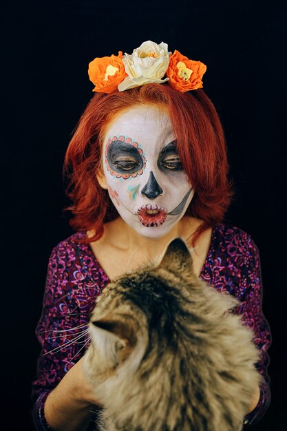 Jonge vrouw in dag van de dode masker schedel gezicht kunst make-up en rood haar spelen met kat op donkere rug...