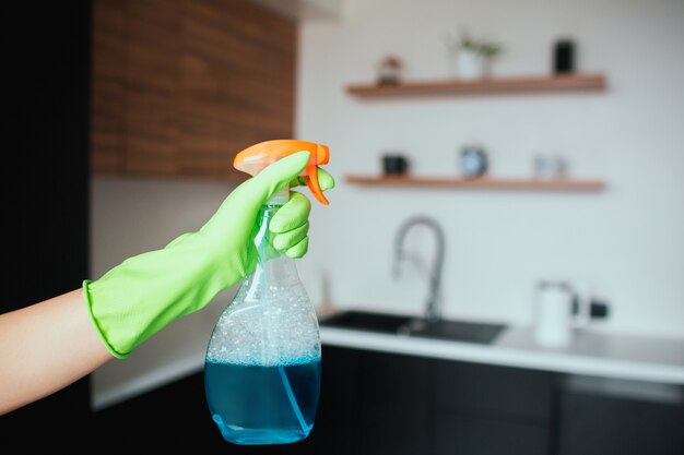 Jonge vrouw in de keuken tijdens de quarantaine. Het beeld van wijfje dient groene het vensternevel van de handschoengreep in. Blauwe vloeistof in de fles. Onscherpe achtergrond.