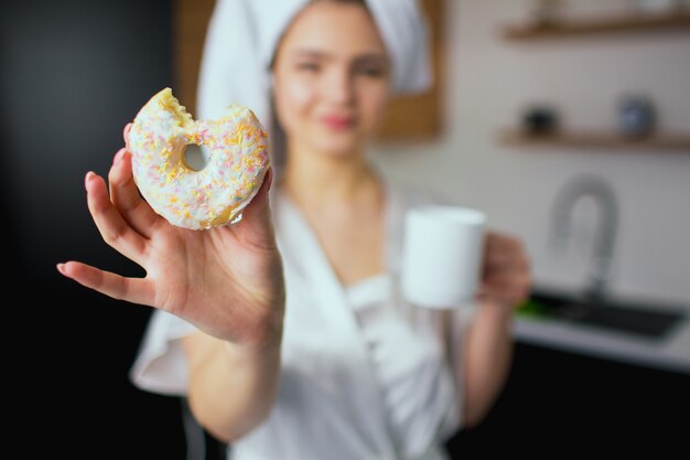 Jonge vrouw in de keuken tijdens de quarantaine. Vaag beeld van meisje na het baden in witte handdoek en robe die stuk van doughnut in hand houden. Kopje drinken in een andere.