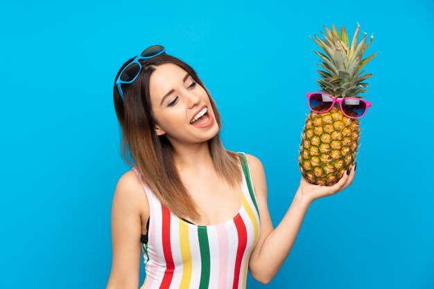 Jonge vrouw in de zomervakantie die over blauw een ananas met zonnebril houdt