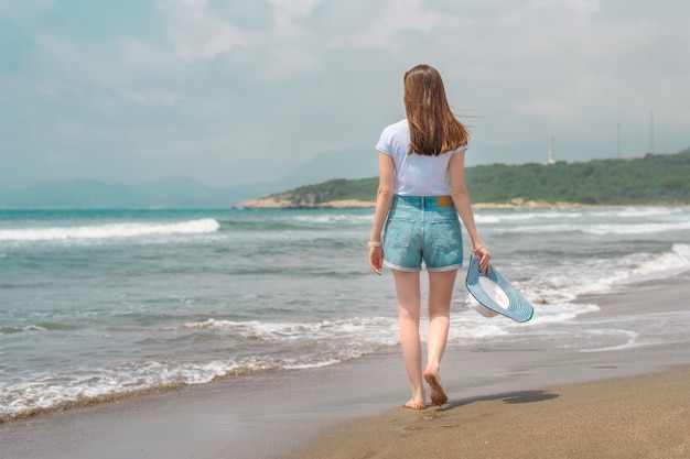 Jonge vrouw in denim shorts en wit t-shirt wandelen langs de middellandse zeekust