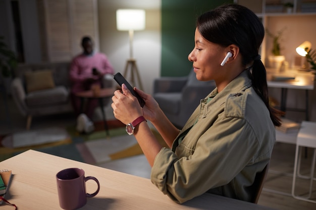 Jonge vrouw in draadloze koptelefoon online werken op haar mobiele telefoon aan de tafel op kantoor