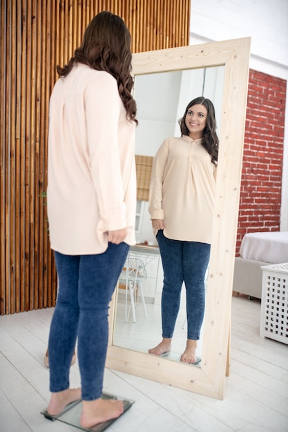 Jonge vrouw in een beige blouse die zich op een weger bevindt en naar zichzelf kijkt