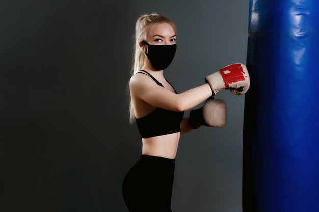 Jonge vrouw in een beschermend masker raakt een bokszak. Beschermende maskers tegen virusinfectie. Trainen tijdens quarantaine in de sportschool