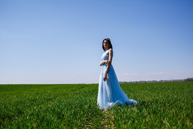 Jonge vrouw in een blauwe lange jurk op een achtergrond van groen veld. Modeportret van een mooi meisje met een glimlach op haar gezicht