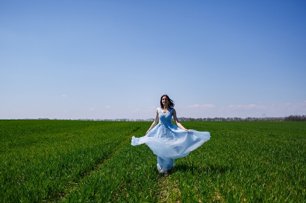 Jonge vrouw in een blauwe lange jurk op een achtergrond van groen veld. Modeportret van een mooi meisje met een glimlach op haar gezicht