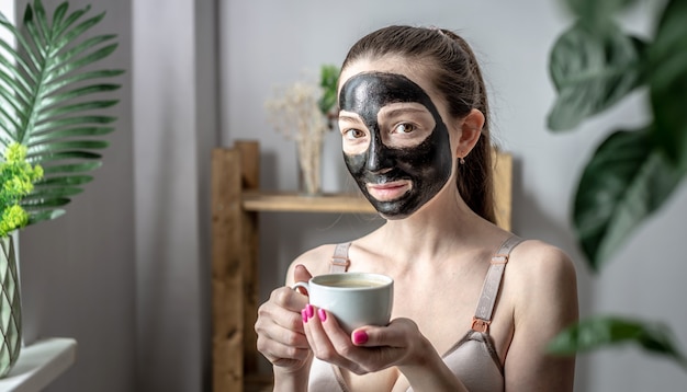 Jonge vrouw in een cosmetisch zwart gezichtsmasker met kopje koffie