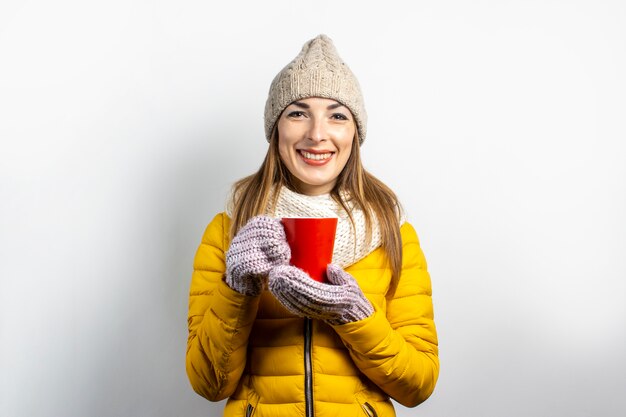 Jonge vrouw in een gele jas en hoed houdt een glas koffie of thee op een lichte achtergrond.