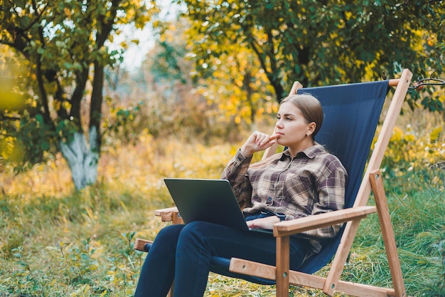 Jonge vrouw in een geruit overhemd zittend in een stoel buiten in een tuin en werkend op een laptop externe werk vrouwelijke freelancer