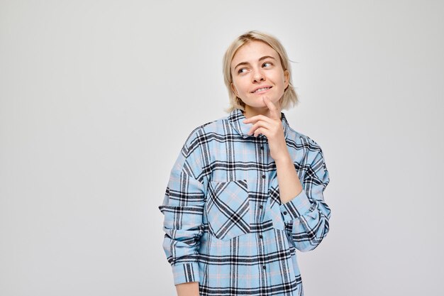 Jonge vrouw in een geruite hemd die onzeker kijkt met de hand op de kin tegen een grijze achtergrond