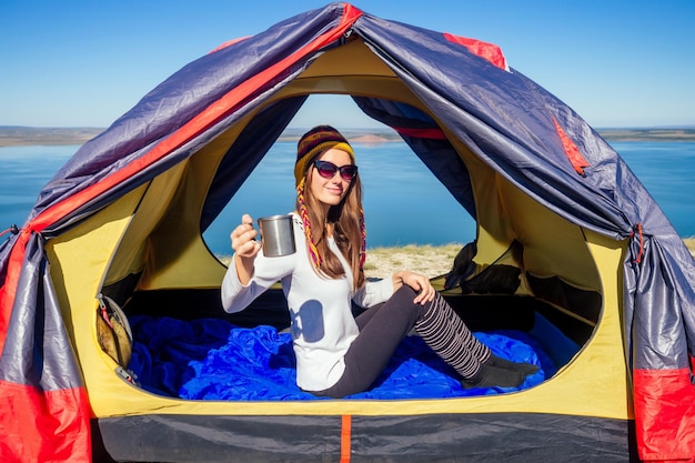 Jonge vrouw in een grappige hoed uit Nepal zittend in een blauwe slaapzak in de tent met een thermos mok en het drinken van koffiekopje thee achtergrond landschap meer. goedemorgen op reis