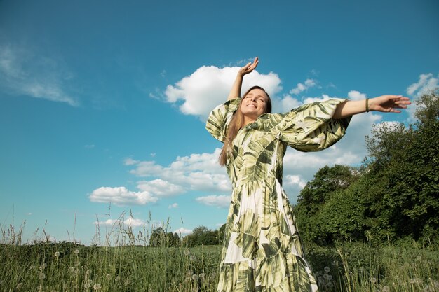 Jonge vrouw in een groen veld met uitgestrekte handen naar de blauwe lucht, geluk