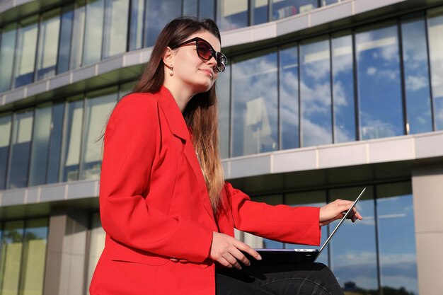 Jonge vrouw in een jas die met een laptop werkt tegen de achtergrond van een kantoorgebouw