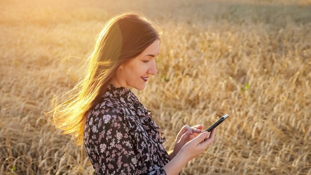 Jonge vrouw in een jurk kijkt naar de telefoon, staat in een tarweveld, zonlicht in haar haar.