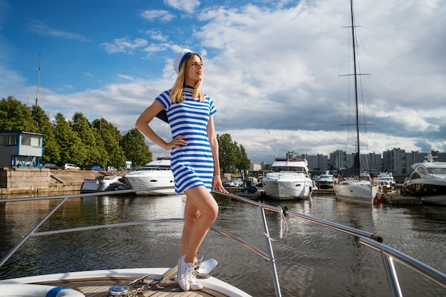 Jonge vrouw in een korte jurk met blauwe strepen en een pet, op een jacht, poserend op een zonnige dag