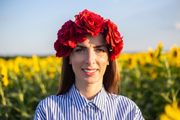 Jonge vrouw in een krans van rode bloemen op een zonnebloemveld bij zonsondergang.