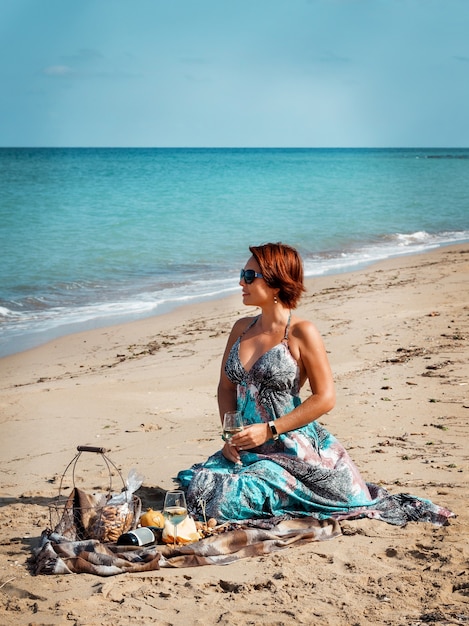 Jonge vrouw in een lange jurk zittend op het strand en wijn drinken