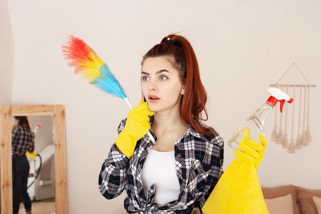 Jonge vrouw in een overhemd en rubberhandschoenen die in het huis schoonmaken en over telefoon spreken