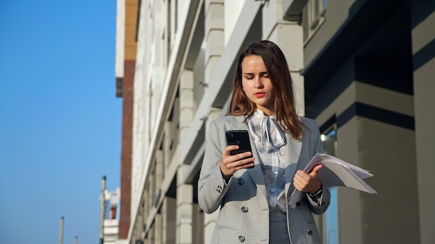 Jonge vrouw in een pak met een telefoon en documenten die over straat lopen.