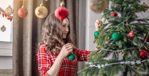 Jonge vrouw in een rode jurk versiert de kerstboom met speelgoed en ballen. Concept van voorbereiding op het nieuwe jaar, gezellige sfeer en feeststemming.