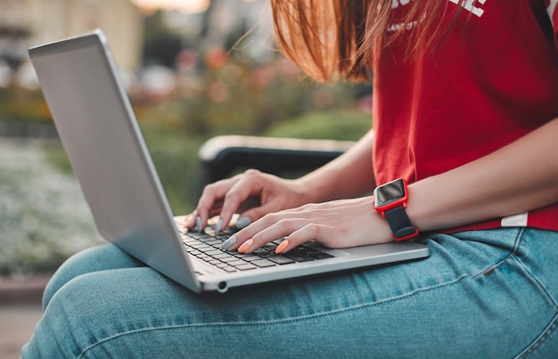 Jonge vrouw in een rood T-shirt en spijkerbroek met een slimme klok zit op een bankje met een laptop op haar schoot a