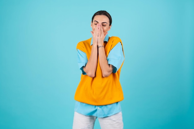 Jonge vrouw in een shirt en een oranje vest op een turkooizen achtergrond lacht vrolijk en bedekt haar gezicht met haar handen